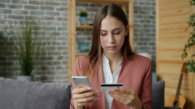 A young woman in a pink blazer shops online using a smartphone and credit card indoors. So aktivieren Sie TWINT in einem Schweizer Onlineshop