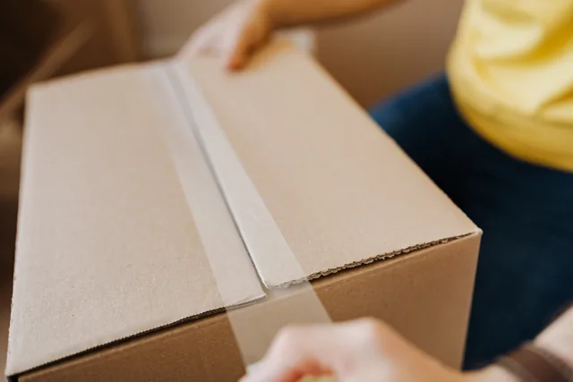 Crop anonymous courier in yellow shirt packing purchase and sealing carton box with tape before deliver. Versand und Retouren für Online-Einkäufe in der Schweiz organisieren
