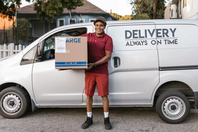 A courier in a maroon uniform smiling with a package in front of a delivery van. Communiquer les frais de livraison et les délais de livraison