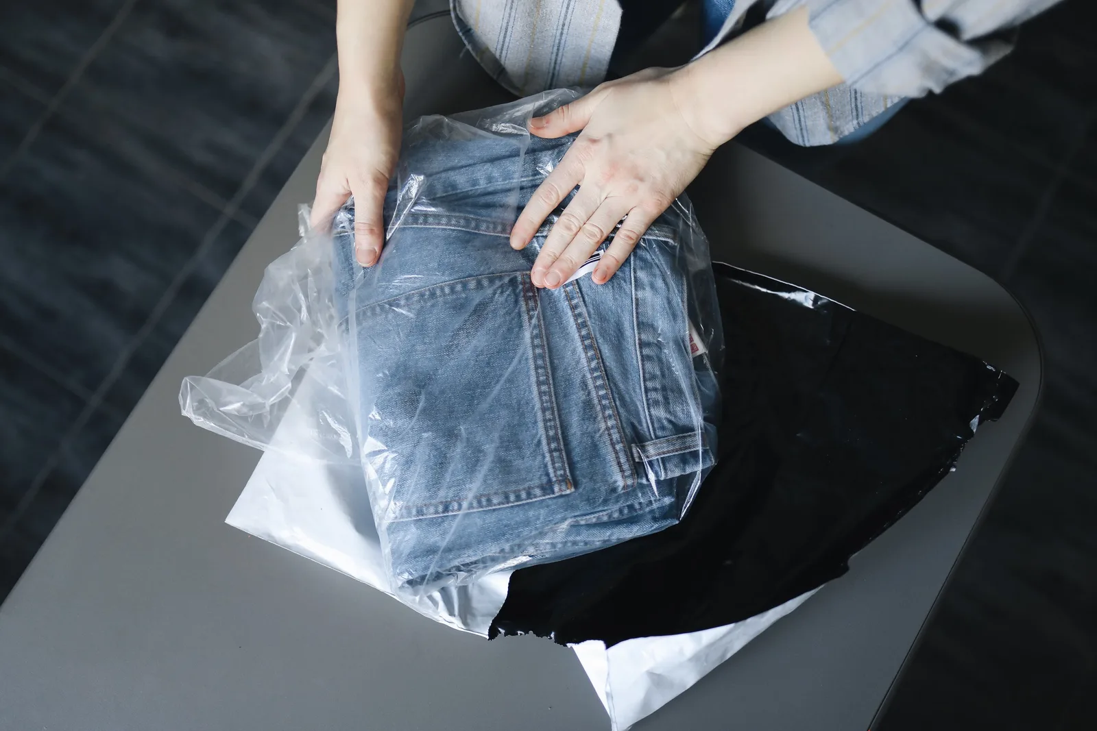Woman carefully packs blue jeans into a plastic bag on a dark surface.